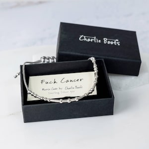 May include: A silver Morse code bracelet spelling "Fuck Cancer" is presented in an open black box. The box contains a card with the message and the brand name "Charlie Boots". The bracelet is strung on a grey cord.
