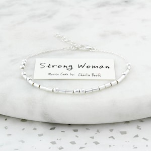 May include: A silver bracelet with a Morse code design. The bracelet features a series of small, cylindrical beads and round beads. A small card with the words "Strong Woman" and "Morse Code by: Charlie Boots" is included.