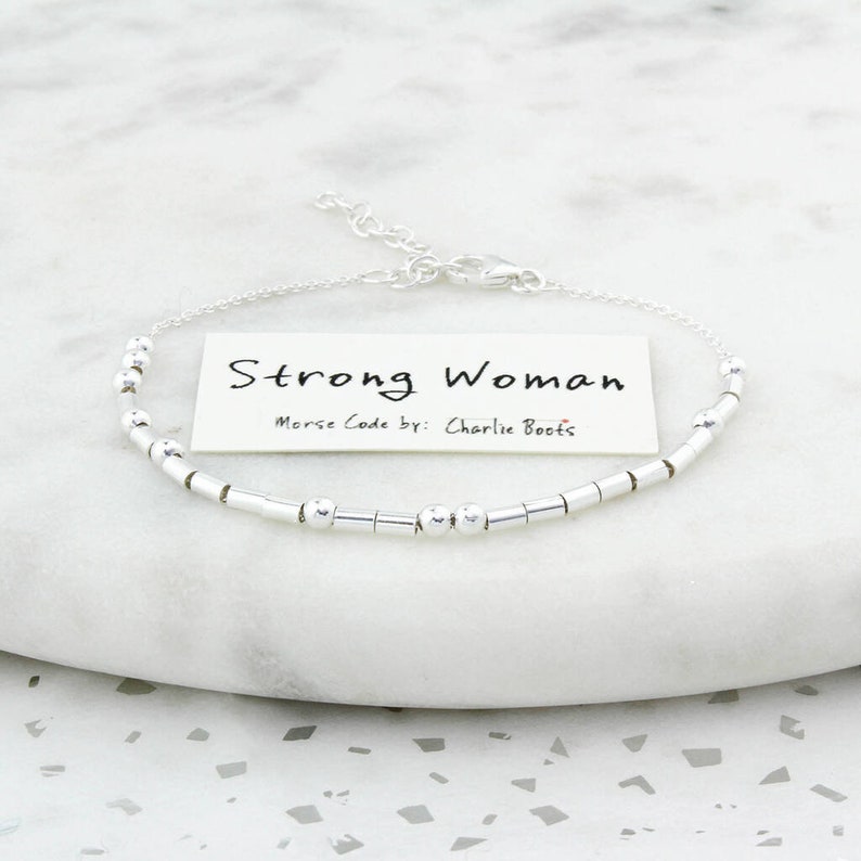 May include: A silver bracelet with a Morse code design. The bracelet features a series of small, cylindrical beads and round beads. A small card with the words "Strong Woman" and "Morse Code by: Charlie Boots" is included.