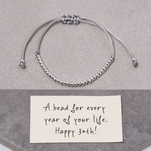 Op de afbeelding: Een zilveren kralen armband op een grijze achtergrond met een kaart waarop staat: "Een kraal voor elk jaar van je leven. Gefeliciteerd met je 30ste verjaardag!"