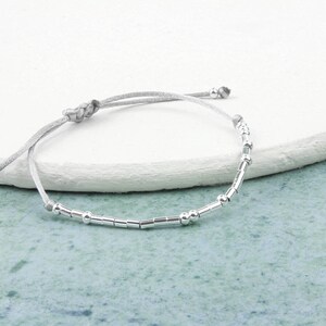 May include: A silver beaded bracelet with a gray cord. The bracelet has small round beads and longer, rectangular beads.