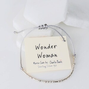 Puede incluir: Un brazalete de plata con un cord&oacute;n gris y una etiqueta que dice "Wonder Woman" en cursiva. La etiqueta tambi&eacute;n dice "Morse Code by: Charlie Boots" y "Sterling Silver 925".
