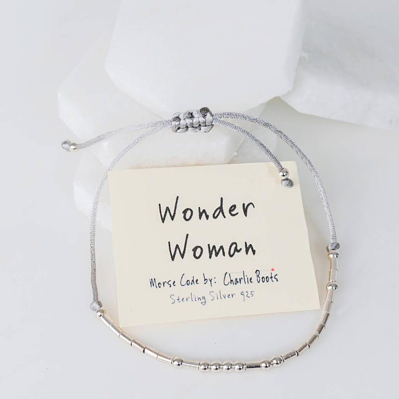 Puede incluir: Un brazalete de plata con un cord&oacute;n gris y una etiqueta que dice "Wonder Woman" en cursiva. La etiqueta tambi&eacute;n dice "Morse Code by: Charlie Boots" y "Sterling Silver 925".