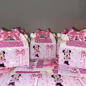 Puede incluir: Un conjunto de cajas de regalo de cartón rosa claro con un gráfico de un ratón de dibujos animados con vestido de lunares, un número uno blanco grande y el nombre EMMA en letras rosas. Cada caja tiene un lazo de satén rosa brillante.