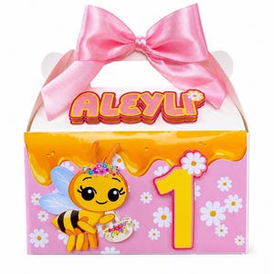 Puede incluir: Una caja blanca con un lazo de satén rosa en la parte superior, con el nombre ALEYLI en letras amarillas. El frente muestra una abeja de dibujos animados, un número 1 amarillo y margaritas blancas sobre un fondo rosa con diseño de miel.