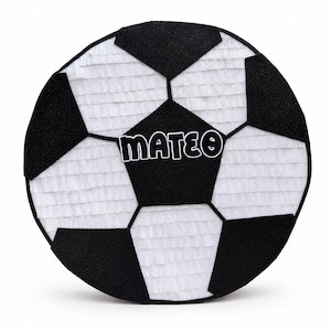 May include: A round soccer ball pinata featuring a classic black and white geometric pattern. The surface is covered in layered fringe paper. A central black pentagon displays the name MATEO in bold white letters with a black outline.