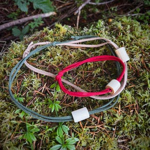 May include: Three handmade bracelets with natural wooden beads. One is red, another is olive green, and the third is tan. The bracelets are displayed on a bed of green moss and forest floor.