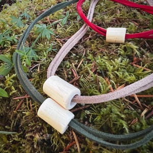 May include: Close-up of three leather cord necklaces with wooden cylindrical beads. One necklace is red, one is tan, and one is green. The necklaces are displayed on a bed of green moss and foliage.