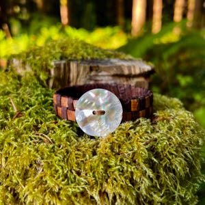 May include: A brown and tan woven leather bracelet with a large, iridescent button. The button has a pearlescent finish. The bracelet is displayed on a bed of green moss. The background includes a wooden stump and natural foliage.