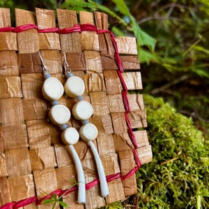 May include: A pair of handmade earrings with a unique design. Each earring features four off-white, circular beads connected by small gray beads and a silver hook. The earrings hang from a woven background of brown and red.