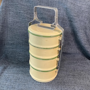 May include: A four-tiered cylindrical food carrier made of cream-colored metal with thin green rims on each stackable container. The unit features a silver-toned metal handle frame that locks the stacked compartments securely in place.