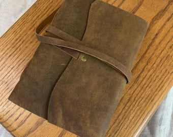 May include: A brown leather journal with a wrap-around strap closure. The journal is rectangular and has a textured surface. The strap is secured with a brass-colored button. The journal is sitting on a wooden surface.
