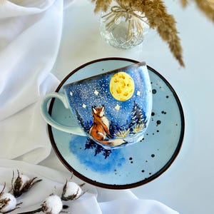 Może przedstawiać: Jasnoniebieski ceramiczny kubek ze spodkiem, ozdobiony ręcznie malowaną sceną przedstawiającą lisa siedzącego w zaśnieżonym lesie pod jasnym, żółtym pełnym księżycem i rozgwieżdżonym niebem. Spodek ma nakrapiane niebieskie wykończenie z brzegiem.