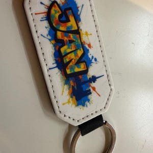 Könnte beinhalten: Weißer, rechteckiger Schlüsselanhänger mit dem Wort "JAN" in farbenfrohen Graffiti-Buchstaben. Das Design umfasst blaue, gelbe, orangefarbene und rote Farbspritzer. Ein schwarzer Riemen verbindet den Schlüsselanhänger mit einem silbernen Schlüsselring.