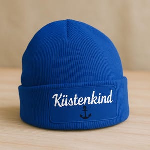 Könnte beinhalten: Eine leuchtend blaue Beanie-Mütze mit umgeschlagenem Saum. Die Mütze hat einen rechteckigen Aufnäher mit dem weißen Text "Küstenkind" und einem schwarzen Ankersymbol. Die Mütze ist aus weichem Strickmaterial gefertigt.