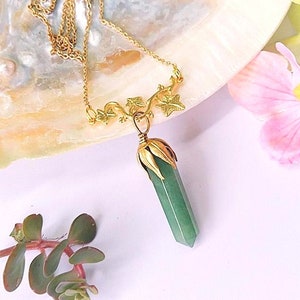 May include: A gold necklace with a green gemstone pendant and a delicate ivy design. The necklace is displayed on a white seashell with pink flowers.