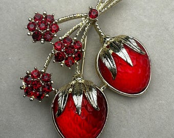 May include: A gold-toned brooch featuring a cluster of red rhinestone flowers and two red enameled strawberry pendants with gold leaf accents. The brooch has a textured gold stem and a pin closure.