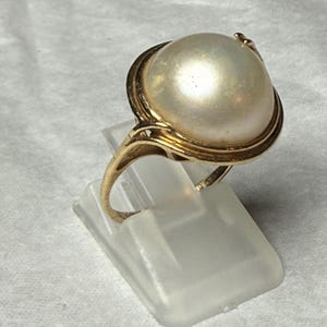 May include: A gold ring featuring a large, round, iridescent white pearl set in a polished gold band. The pearl has a smooth, lustrous surface and is cradled by a delicate gold setting that wraps around the base of the gemstone, creating a classic look.