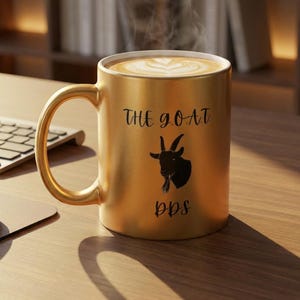May include: A gold-coloured coffee mug with the text "THE GOAT" and a goat silhouette, with the letters "bbs" below. The mug is filled with coffee and has a latte art design on top. Steam rises from the coffee.