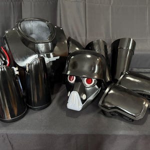 Puede incluir: Un conjunto de piezas de armadura negras que incluye una placa de pecho, un casco con lentes oculares rojas y una rejilla bucal blanca, hombreras, protectores de brazo y espinilleras. La armadura tiene un acabado brillante y correas.