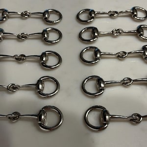 May include: A set of ten silver-toned metal horse bit hardware pieces arranged in two columns of five. Each piece features a central bar connected to two circular rings on either end, designed for use as decorative fasteners or equestrian-style accents.