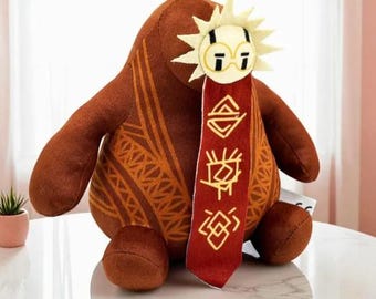 Peut inclure: Peluche marron avec un ornement en forme de soleil et un ruban rouge orné de symboles dorés. Le jouet présente un motif sur son corps et est posé sur une surface blanche. Fabriqué en matière douce.