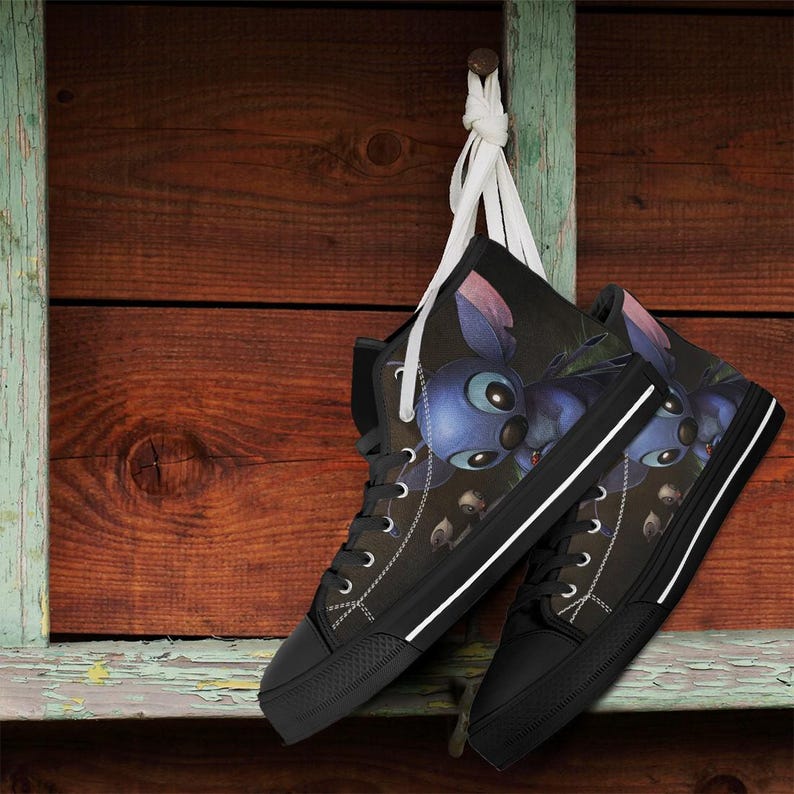 May include: Black high-top sneakers featuring a blue cartoon character design. The shoes have black soles, white laces, and a white stripe along the side. The sneakers are hanging against a wooden background.