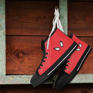 May include: Red high-top sneakers with black soles and laces, featuring white eye designs on the sides. The shoes are hanging against a weathered wooden background, showcasing a unique design.