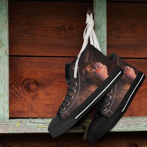 May include: A pair of high-top canvas sneakers with a black sole and laces. The sneakers feature a brown and black graphic print of two cartoon characters. The shoes are hanging against a wooden background.