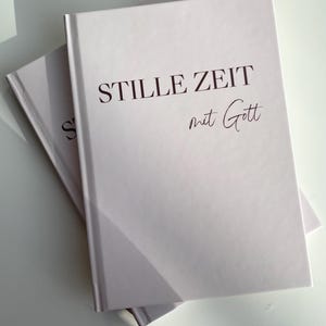 Könnte beinhalten: Zwei hellviolette Hardcover-Bücher liegen auf einer weißen Oberfläche. Auf dem oberen Buch steht in dunkelbrauner Schrift "STILLE ZEIT mit Gott". Die Bücher sind leicht angewinkelt, mit Schatten auf der Oberfläche.