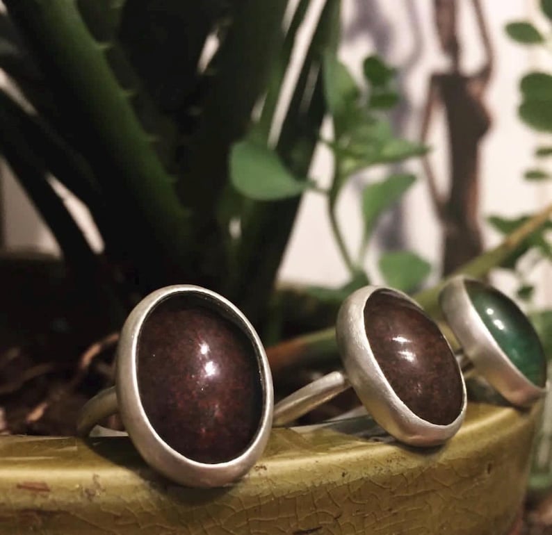 May include: Three rings with oval-shaped stones in shades of brown and green, set in silver bands. The rings are displayed on a green surface, with a blurred background of plants and a silhouette.