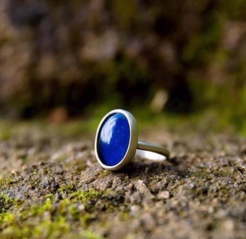 May include: A silver ring with a large, oval, deep blue stone. The ring is set on a natural, earthy surface, with a blurred background of green and brown. The ring's design is simple and elegant.