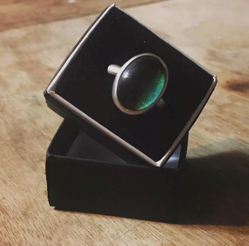 May include: A silver ring with an oval, iridescent stone, displayed in a black ring box. The stone transitions from dark green to a lighter teal color. The ring is set in a simple, polished silver band.