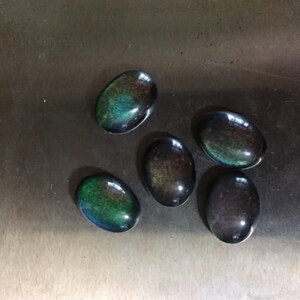 May include: Five oval-shaped cabochon gemstones with a polished, iridescent finish. The stones display a mix of colors, including green, blue, and brown, with a dark base. The gemstones are arranged on a metallic surface.