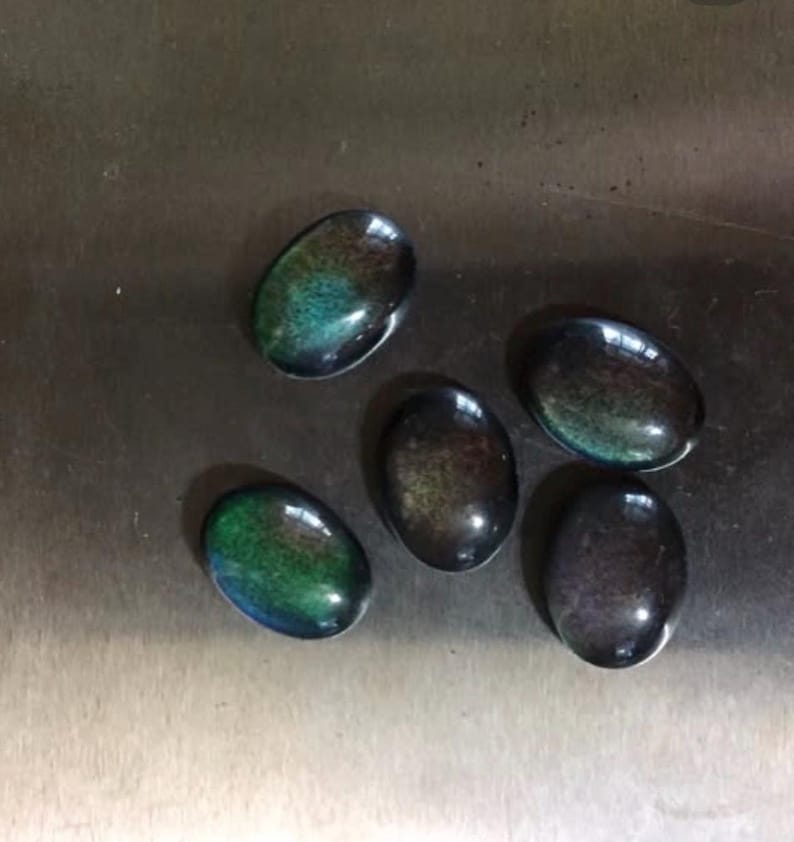 May include: Five oval-shaped cabochon gemstones with a polished, iridescent finish. The stones display a mix of colors, including green, blue, and brown, with a dark base. The gemstones are arranged on a metallic surface.