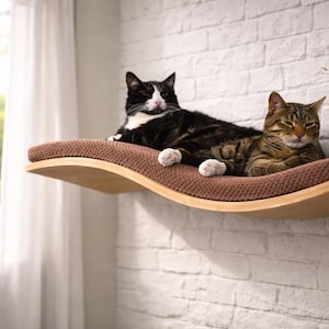 May include: A wooden cat shelf with a brown cushion, mounted on a white brick wall. Two cats, one black and white, the other brown tabby, are resting on the shelf. The shelf has a curved design.