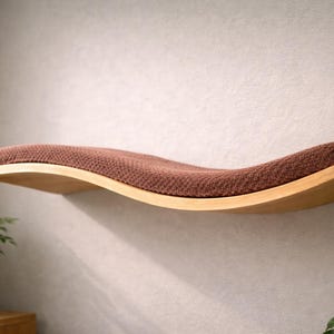 May include: A wooden cat shelf with a curved design, featuring a brown, textured cushion. The shelf is mounted on a light-colored wall, providing a comfortable and stylish resting spot for pets. The shelf is approximately 30 inches long.