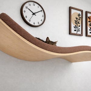 May include: A modern cat shelf in a wave design, made of light wood with a brown, textured cushion. A tabby cat rests on the shelf. A round clock and two framed botanical prints hang on the white wall behind the shelf.