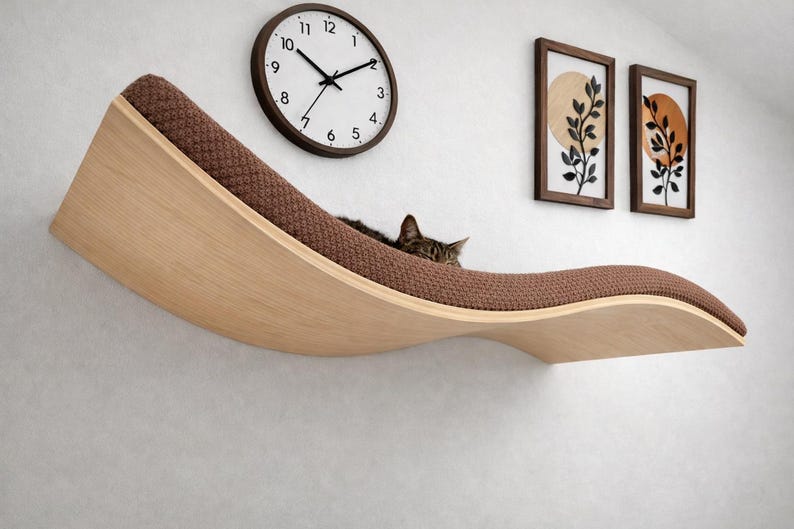 May include: A modern cat shelf in a wave design, made of light wood with a brown, textured cushion. A tabby cat rests on the shelf. A round clock and two framed botanical prints hang on the white wall behind the shelf.