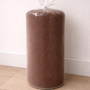 May include: A cylindrical roll of brown fabric, tightly wrapped in clear plastic. The fabric has a textured, woven appearance. The plastic is tied at the top with a twist tie. The roll is standing upright on a light-colored wooden floor.