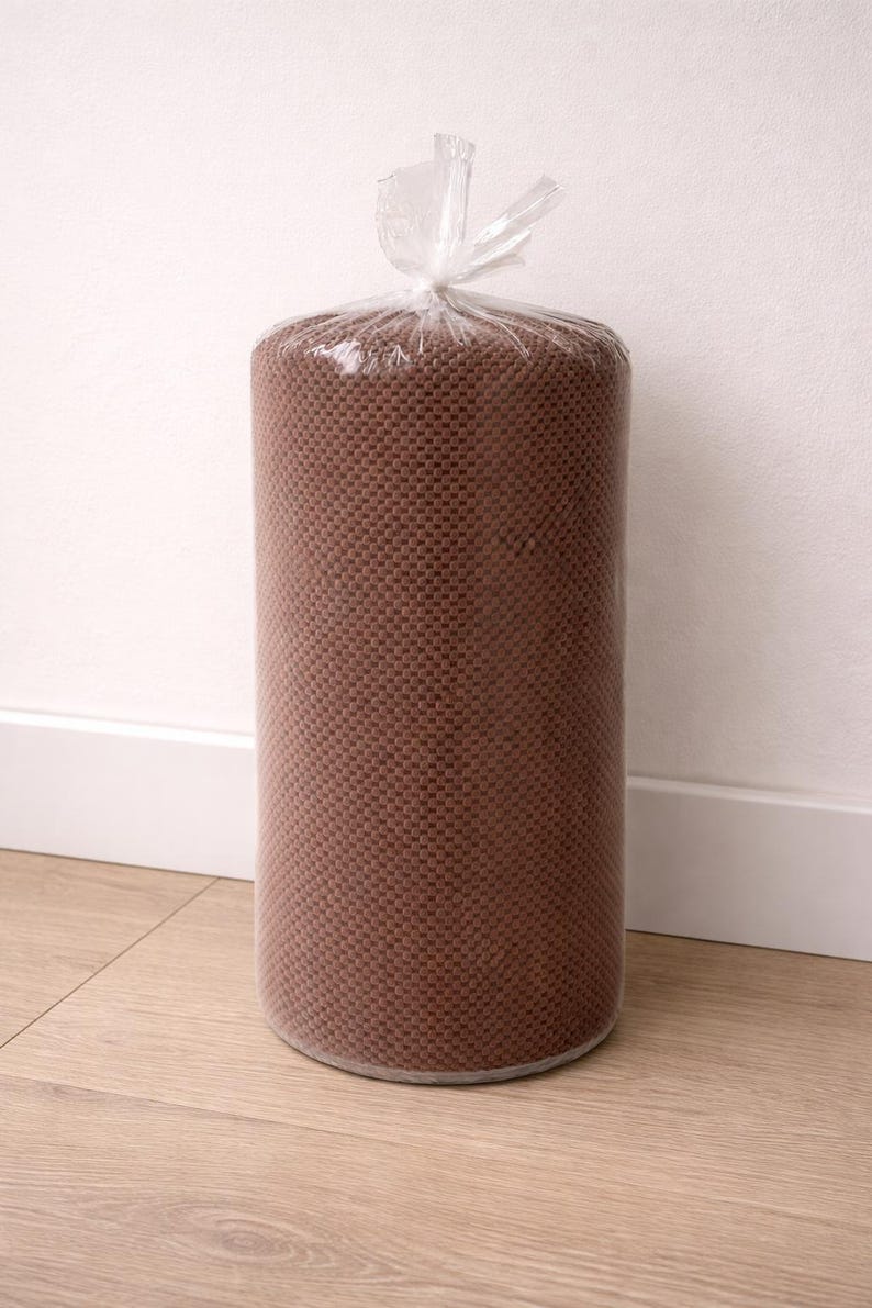 May include: A cylindrical roll of brown fabric, tightly wrapped in clear plastic. The fabric has a textured, woven appearance. The plastic is tied at the top with a twist tie. The roll is standing upright on a light-colored wooden floor.