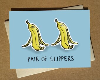 May include: A greeting card with a light blue background featuring two yellow banana peels with sad faces. The text "PAIR OF SLIPPERS" is printed below the banana peels.