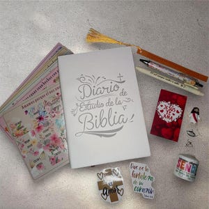 May include: A white Bible study journal with the title "Diario de Estudio de la Biblia" in silver script. The image includes a bookmark, pens, stickers, and a small figurine of Jesus.