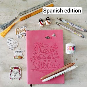 Peut inclure: Un journal d'étude biblique rose avec le titre "Diario de Estudio de la Biblia" est entouré d'accessoires religieux. Sont inclus des stylos, des autocollants, des signets et de petites figurines. Le texte "Spanish edition" est visible.