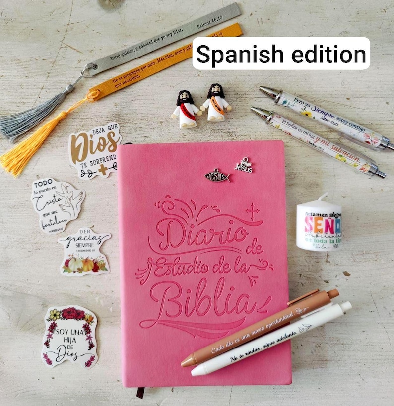 May include: A pink Bible study journal with the title "Diario de Estudio de la Biblia" is surrounded by religious accessories. Included are pens, stickers, bookmarks, and small figurines. The text "Spanish edition" is visible.