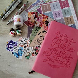 May include: A pink Bible Study Journal with embossed lettering, surrounded by stickers, pens, and bookmarks. The stickers feature inspirational quotes and a butterfly. The pens have floral designs, and the bookmarks have floral patterns and Bible verses.