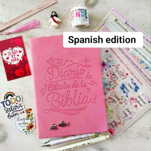 May include: A pink Bible study journal with the title "Diario de Estudio de la Biblia" embossed on the cover. The image includes stickers, pens, a small Jesus figurine, a candle, and a bookmark. The text "Spanish edition" is also visible.