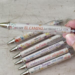 May include: A collection of clear pens with floral designs and silver tips. Each pen features a different inspirational quote in Spanish. The pens are decorated with colorful flowers and text from the Bible.