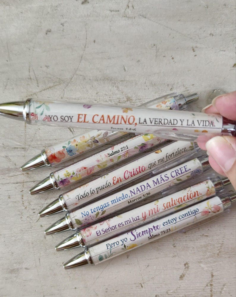 May include: A collection of clear pens with floral designs and silver tips. Each pen features a different inspirational quote in Spanish. The pens are decorated with colorful flowers and text from the Bible.