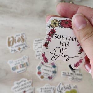 May include: A collection of stickers with floral designs and religious phrases in Spanish. One sticker reads "SOY UNA HIJA DE DIOS" with a floral border. Other stickers feature phrases like "Dios" and "QUE CREE".